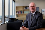 Richard Wynne MP, wearing a suit and tie, sits in an office with windows looking out over a city view.