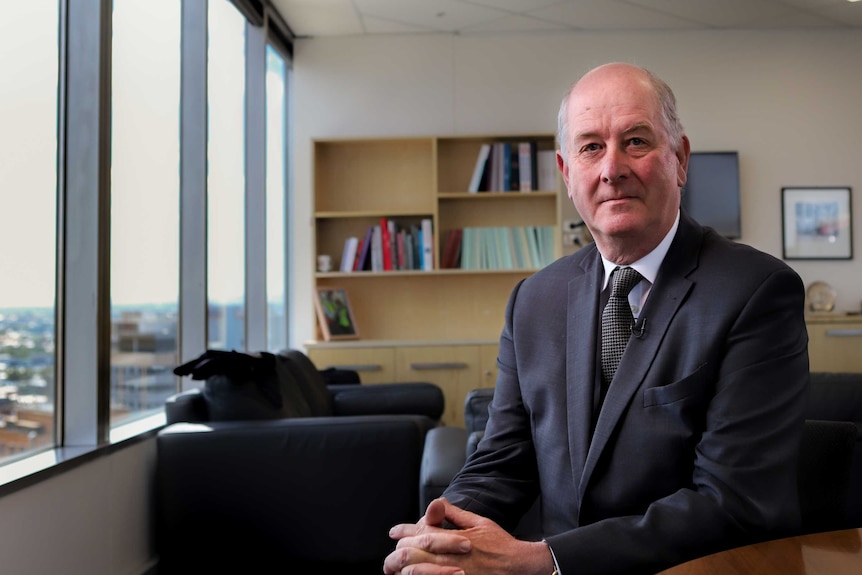 An older man wearing a suit and tie sits in an office with windows looking out over a city view.