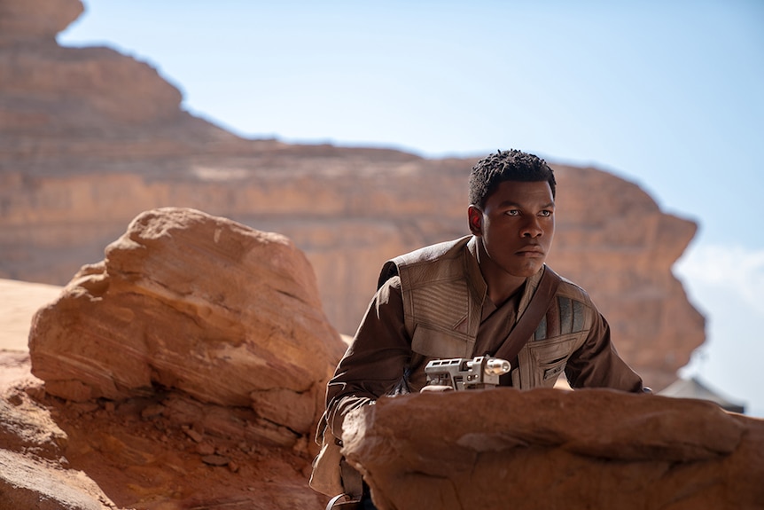 A man with serious and focused expression holds weapon and crouches behind rock in desert landscape on sunny clear day.