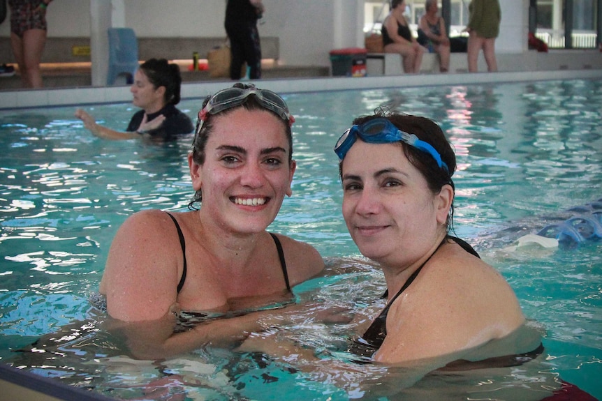 A young woman and her mother, rest on the lane-rope in a pool, smiling at the camera.