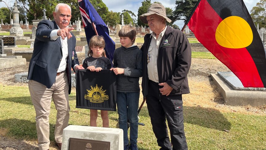 Two men stand next to two children, holding an Australian army flag in front of Australian and Aboriginal flags. 