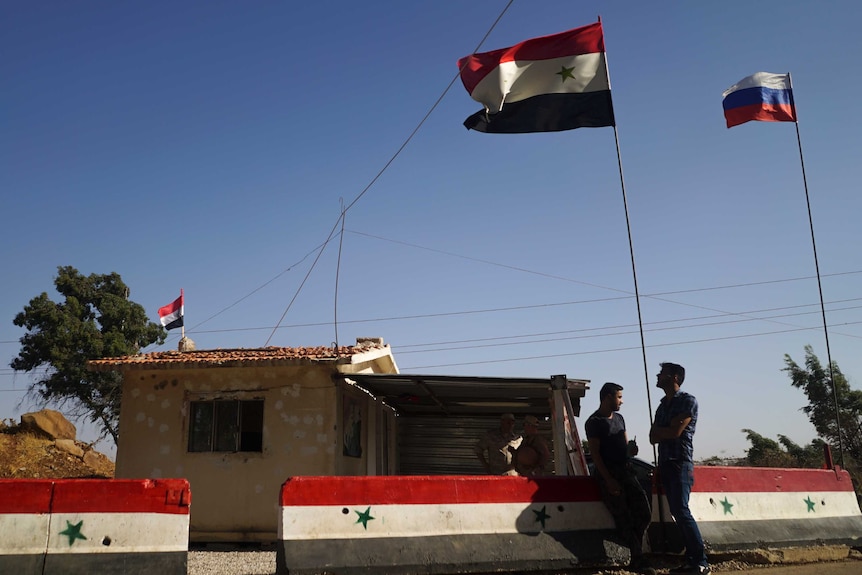 Syrian and Russian flags fly outside a dwelling while people stand near the fence.