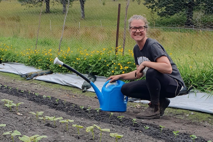 A woman crouches in a paddock next to a row of seedlings with a watering can