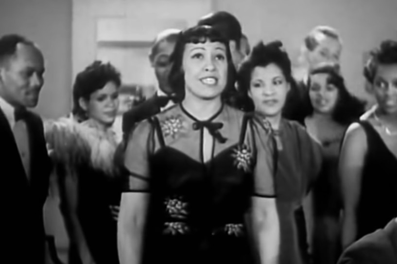 In a black and white scene a woman with fringe and bob hair cut wears dress and stands in small crowd looking up hopefully.