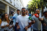 A man wearing sunglasses and a white t-shirt leaves court surrounded by journalists.