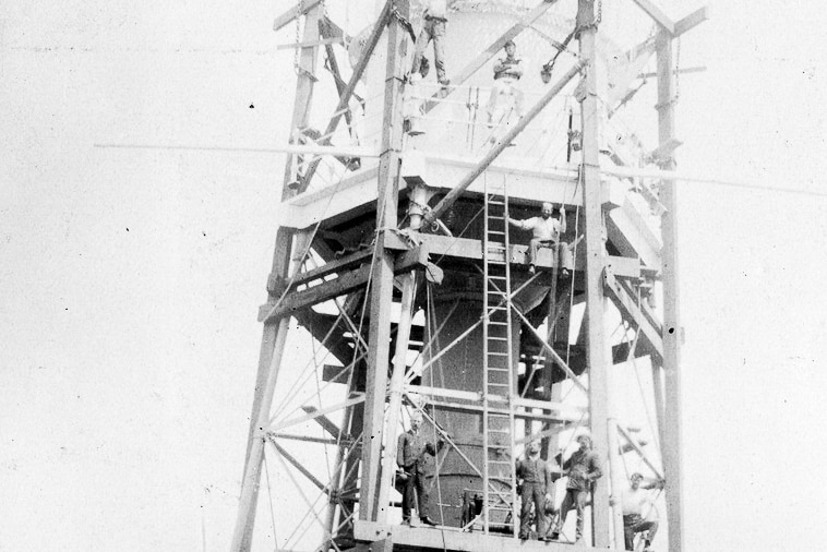 A black and white photograph of several men sitting on rungs of a large lighthouse.