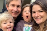 A family selfie of smiling man and woman, holding two small children who are pulling amusing faces.