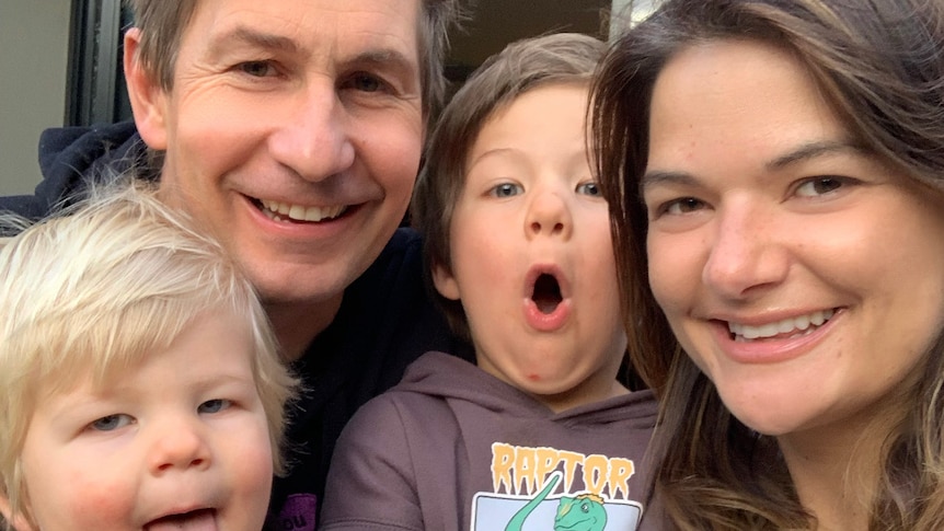 A family selfie of smiling man and woman, holding two small children who are pulling amusing faces.