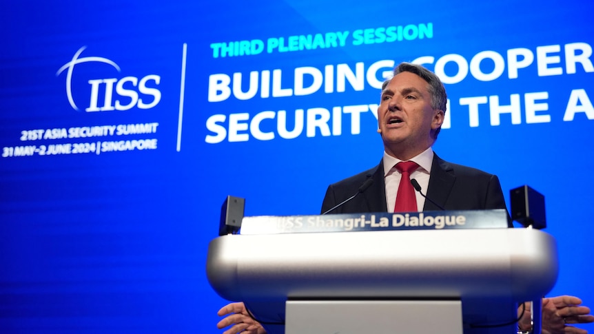 A middle-aged man in a suit with grey hair speaks behind a lectern in front of a blue screen.