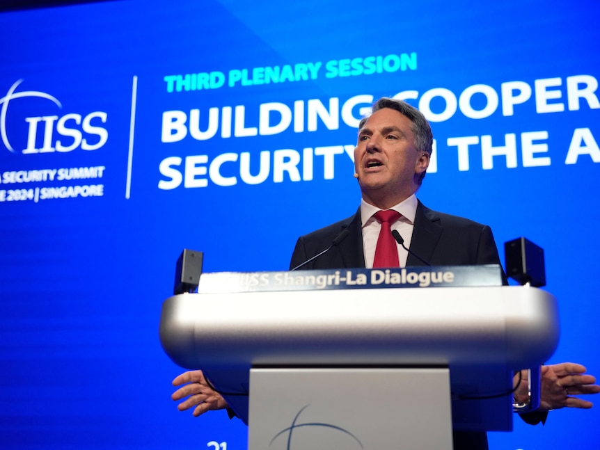 A middle-aged man in a suit with grey hair speaks behind a lectern in front of a blue screen.