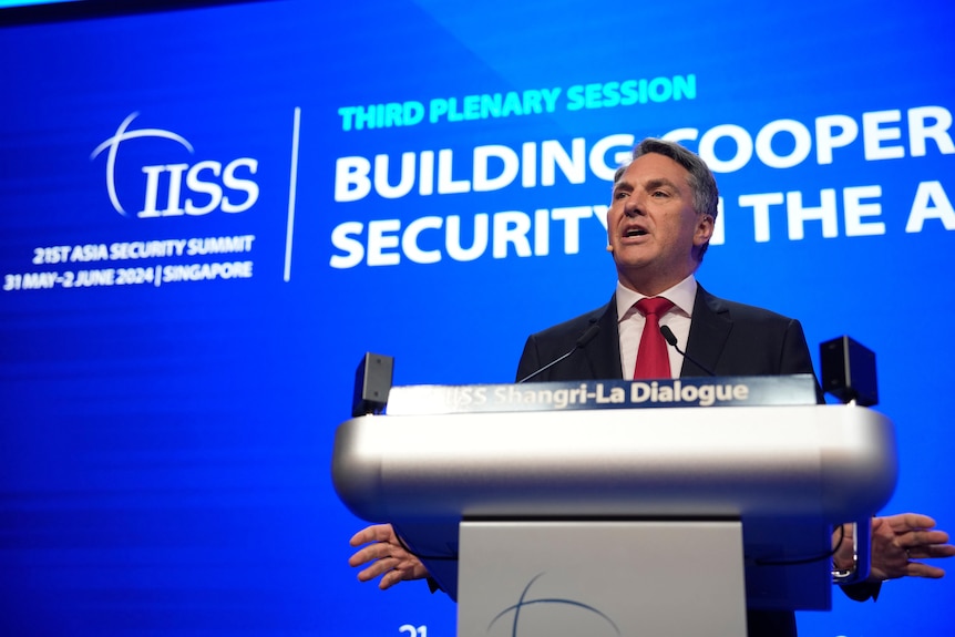 A middle-aged man in a suit with grey hair speaks behind a lectern in front of a blue screen.