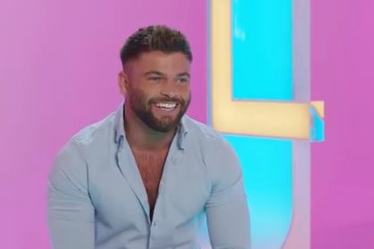 A man wears a light blue shirt and laughs as he sits in front of a pink and blue background with the word love half shown.