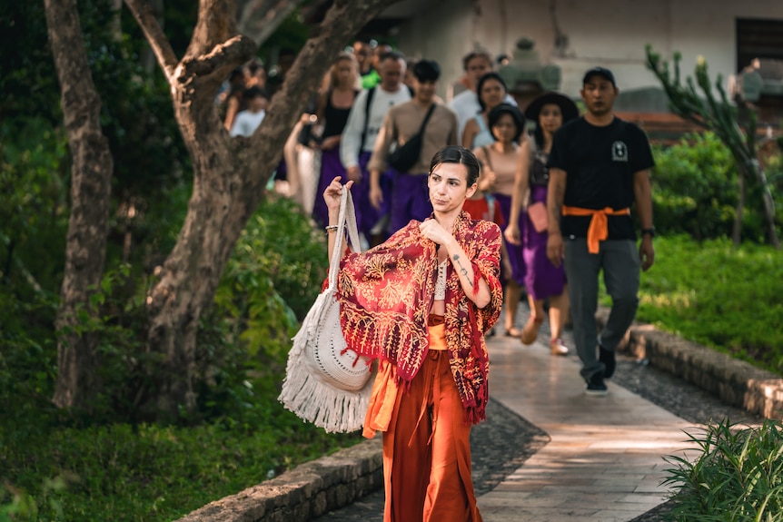 A close up of a woman holding a fringe bag wearing an orange shawl and pants.