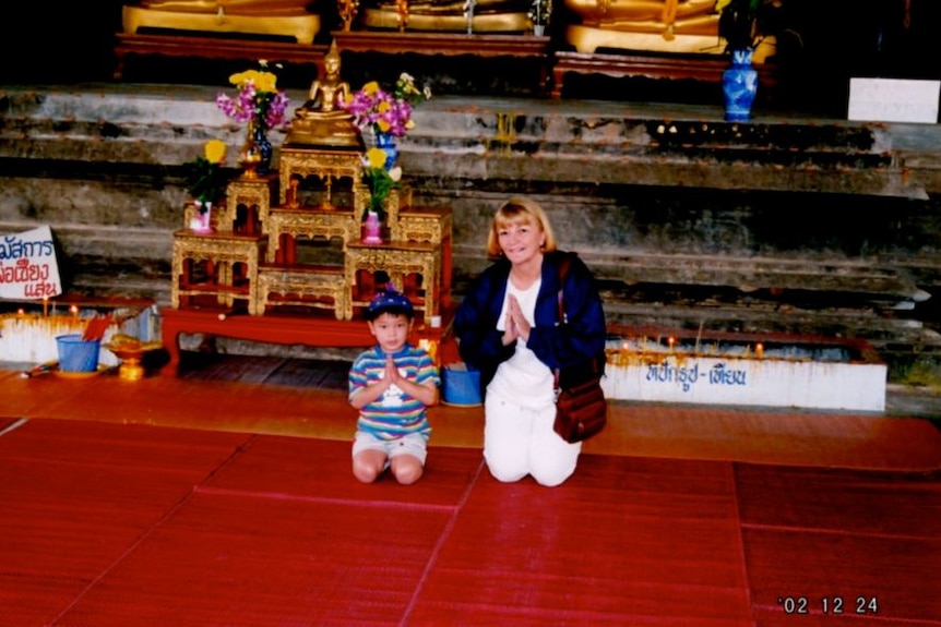 A young Asian child is seen kneeling with hands in prayer next to a woman, his mother. They sit in front of a gold temple.