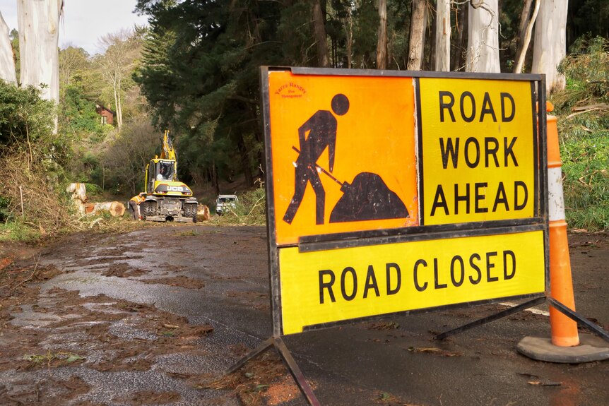 A 'ROAD WORK AHEAD' sign is laid out on a muddy road, with trucks visible further down.