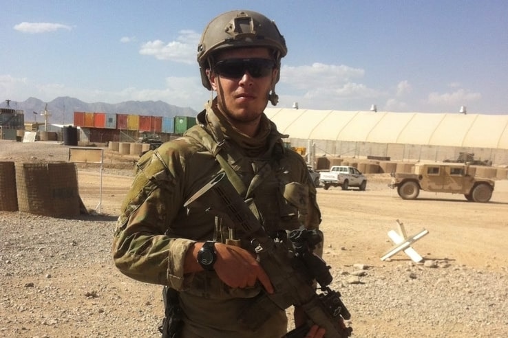 Soldier David Hill stands on gravel holding a gun, wearing army gear and a helmet.