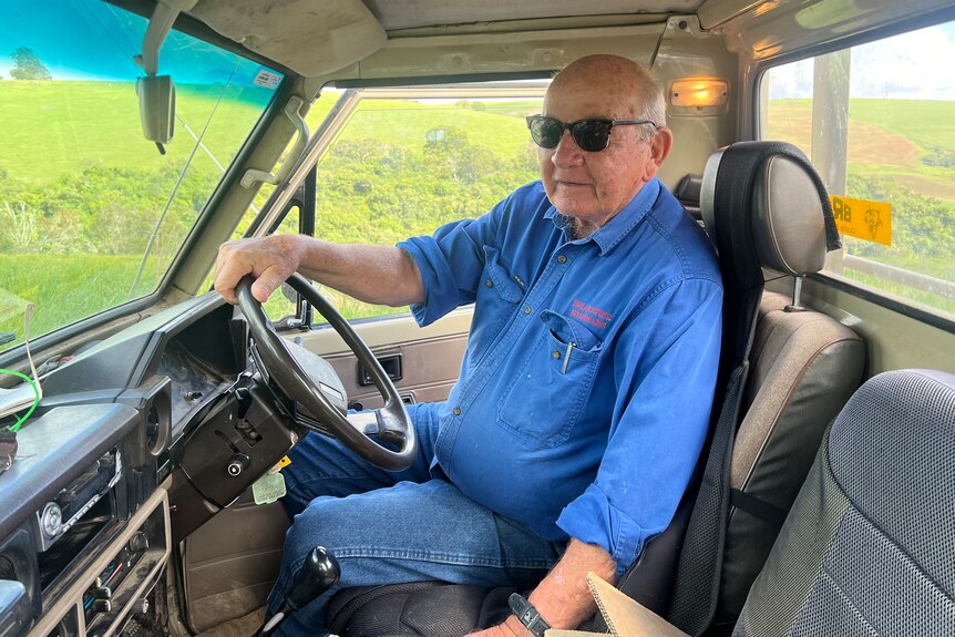 Older man holding steering wheel in stationary car with a blue shirt looking at camera