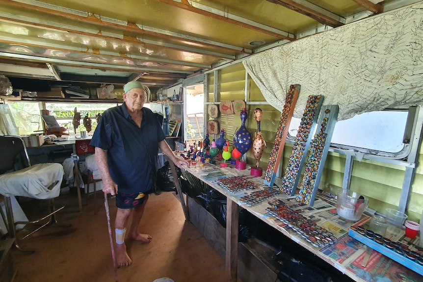 Man leaning on a stick, stands inside a caravan with his art beside him on a table.