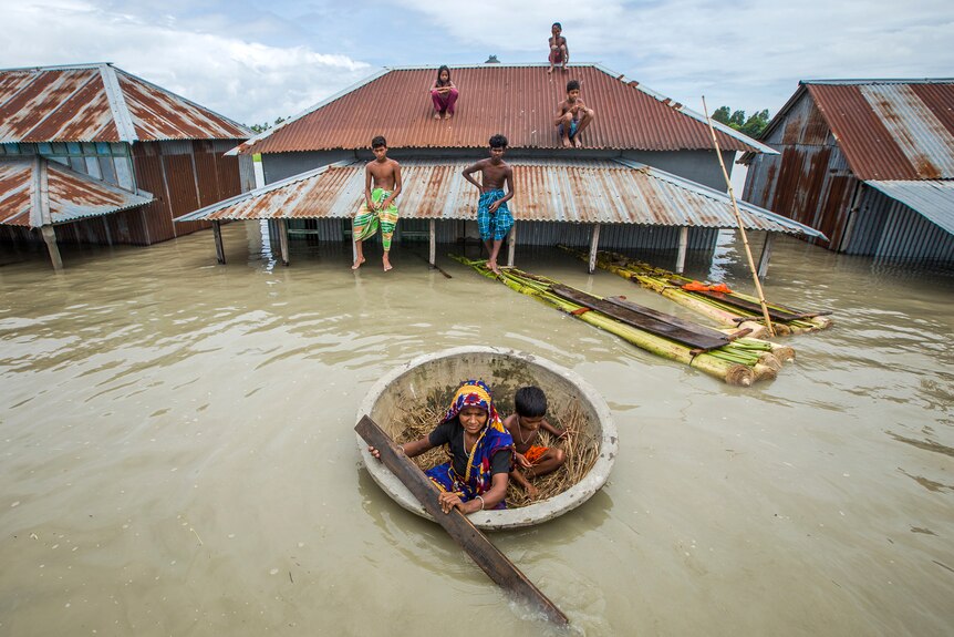 Children sit on the roof of a building that's submerged in brown floodwater, as a woman and child paddle by.