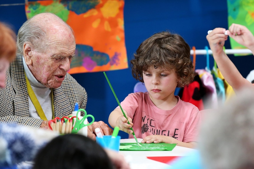 An elderly man in a tweed jacket watches a young child paint on the ABC TV series Old People's Home For 4 Year Olds.