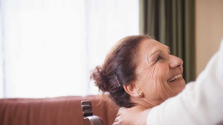 Elderly woman sitting in a chair with carer's hand on her shoulder.