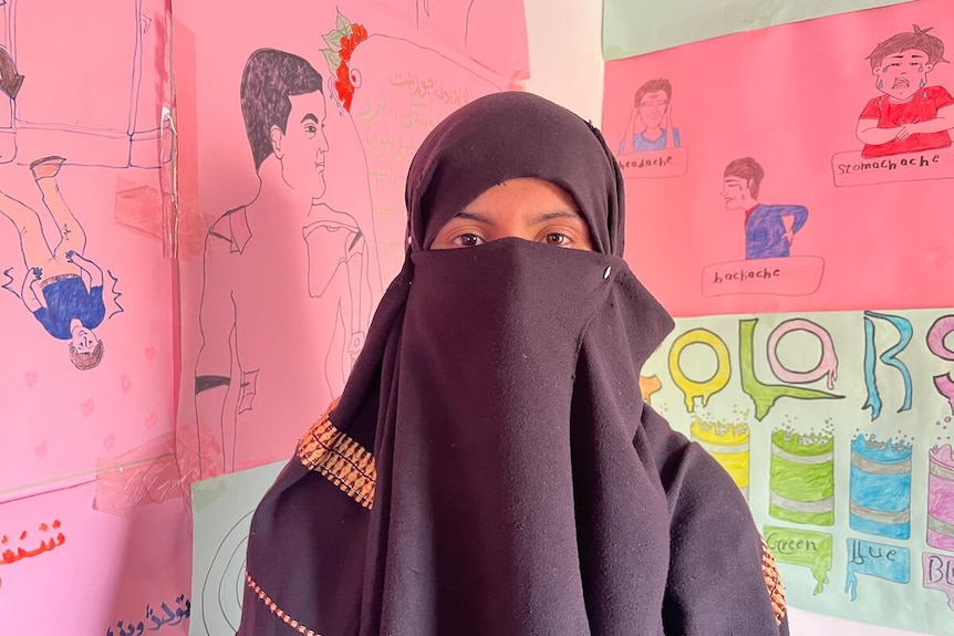 A girl in a black hijab and black niqab covering the lower half of her face with drawings on walls behind her.