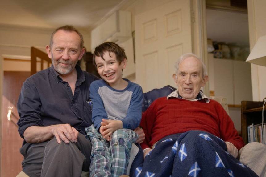 An elderly man, his middle-aged son, and a young boy sit together inside a home.