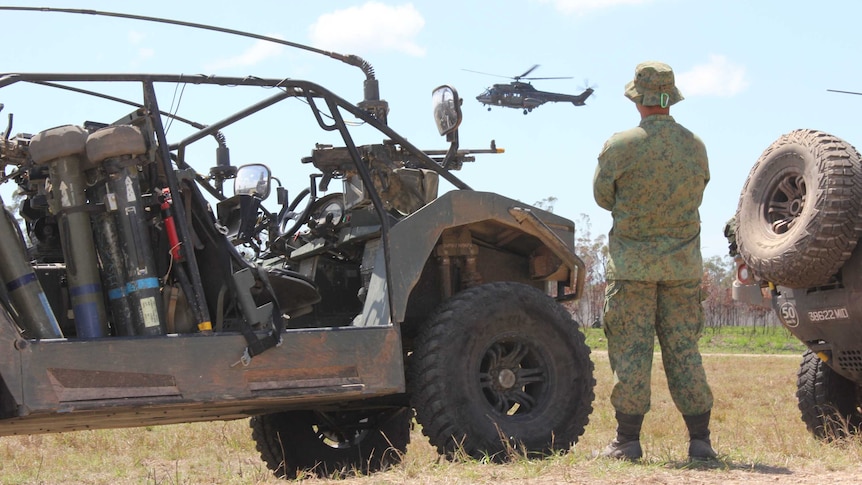 Soldier standing between two vehicles and helicopter overhead