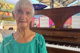 An older woman with close cropped white hair and a green shirt sits in front of a piano.