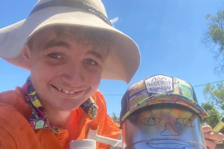 Young man in wide-brimmed hat and orange shirt takes a toothy selfie with a tin-man figure wearing a cap. Ausnew Home Care, NDIS registered provider, My Aged Care