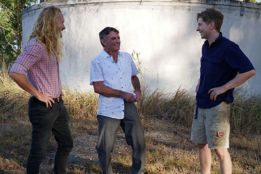 Three men stand laughing with each other in front of a concrete tank on a farm