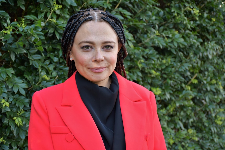 An Aboriginal woman with dreadlocks smiles at the camera in front of a hedge