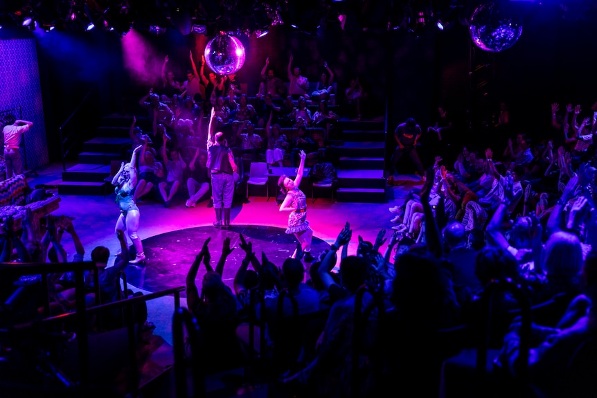 Audience members have their hands above their heads, mimicing moves on-stage. A disco ball and purple lights illuminate.
