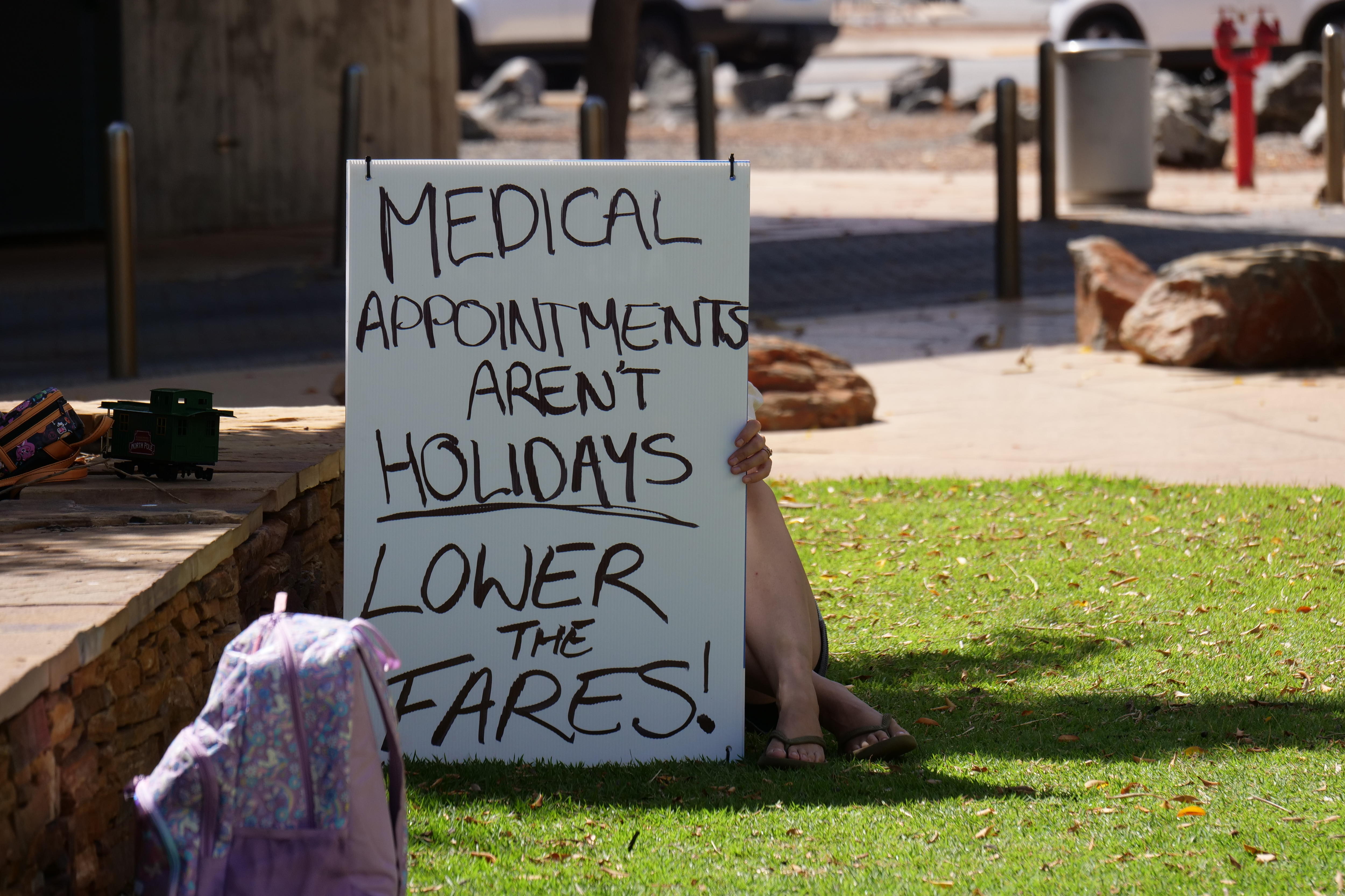 Una persona sentada en el césped y sostiene un cartel pidiendo tarifas aéreas más bajas.