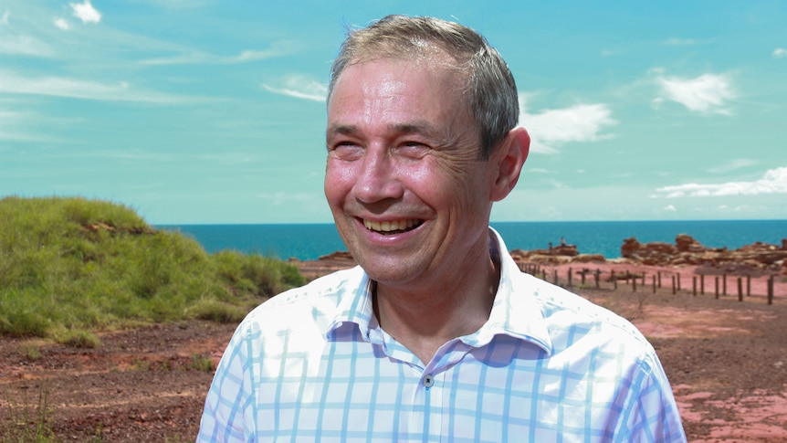 A man smiling with the ocean and red sand behind him.