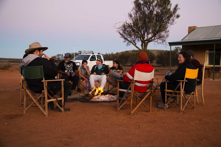 A group of aboriginal and non aboriginal adults and children sit around a camp fire at dusk.
