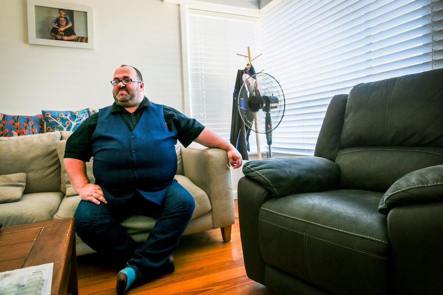 Cameron Bloomfield sits on a couch at home, wearing a vest over a collared shirt.
