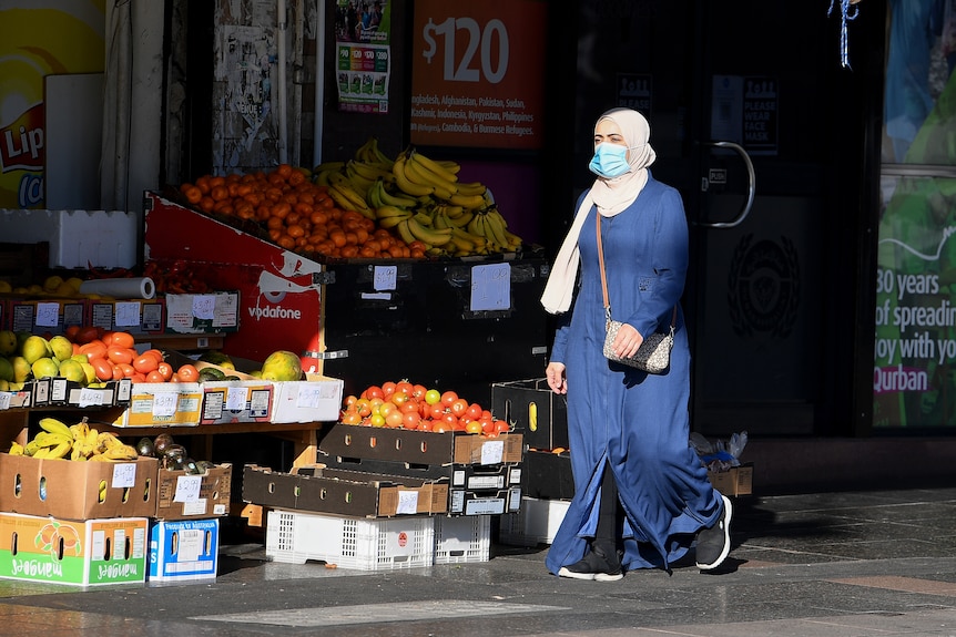 a woman in a hijab and mask walking past a fruit store