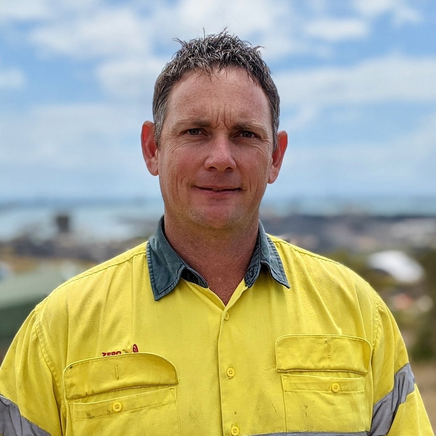 A man in a high vis shirt stands outside on a sunny day, smiling.