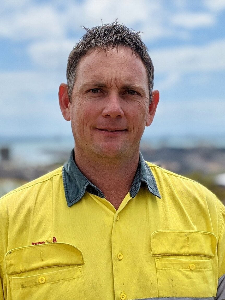 A man in a dirty fluro yellow shirt stands looking at a camera smiling