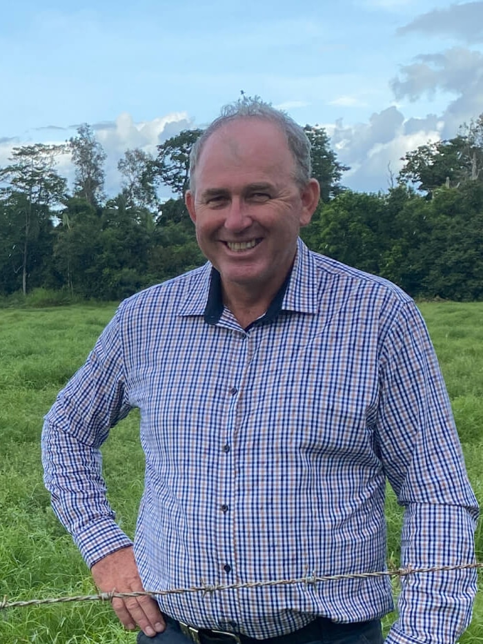 A man wears a check blue shirt smiles at camera with green paddock and cows in background