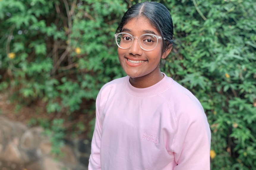 A young girl with brown skin, a pink sweater and glasses smiles for the camera in front of a plant backdrop.