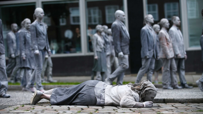 Woman dressed and painted in grey lies on cobble-stoned road as crowd of grey-dressed people walk past