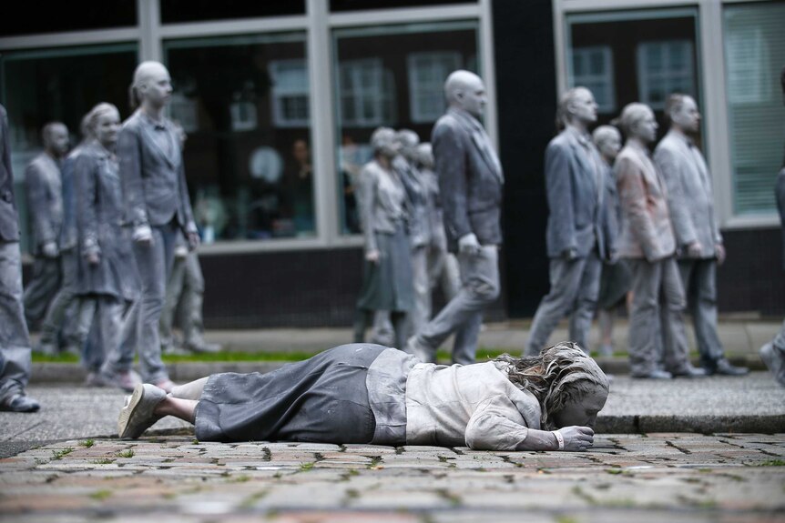 Woman dressed and painted in grey lies on cobble-stoned road as crowd of grey-dressed people walk past