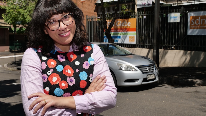 A woman wearing glasses, purple shirt and floral dress smiles with her arms folded