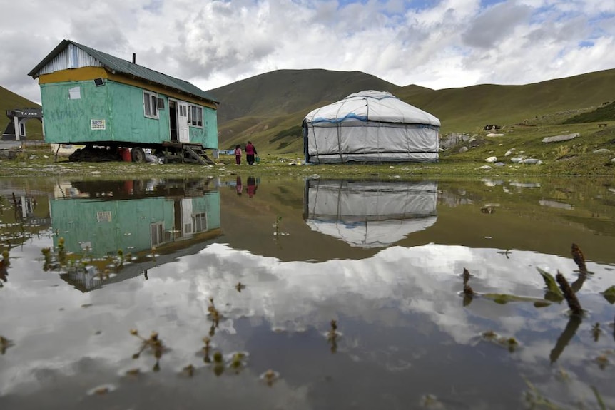 A traditional yurt and a Soviet style temporary house are reflected in a lake in a mountain pasture