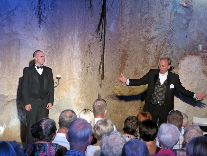 Two men singing, wearing tuxedos. Limestone walls in background and backs of audience heads in foreground