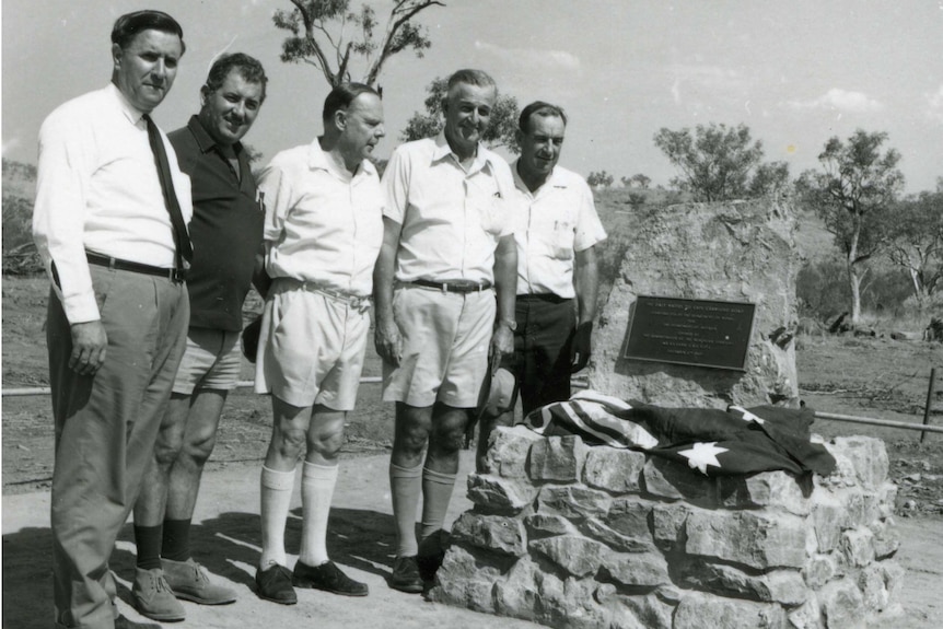 Monochrome of five men posing beside a cairn with plaque. Two men have shorts and long socks.