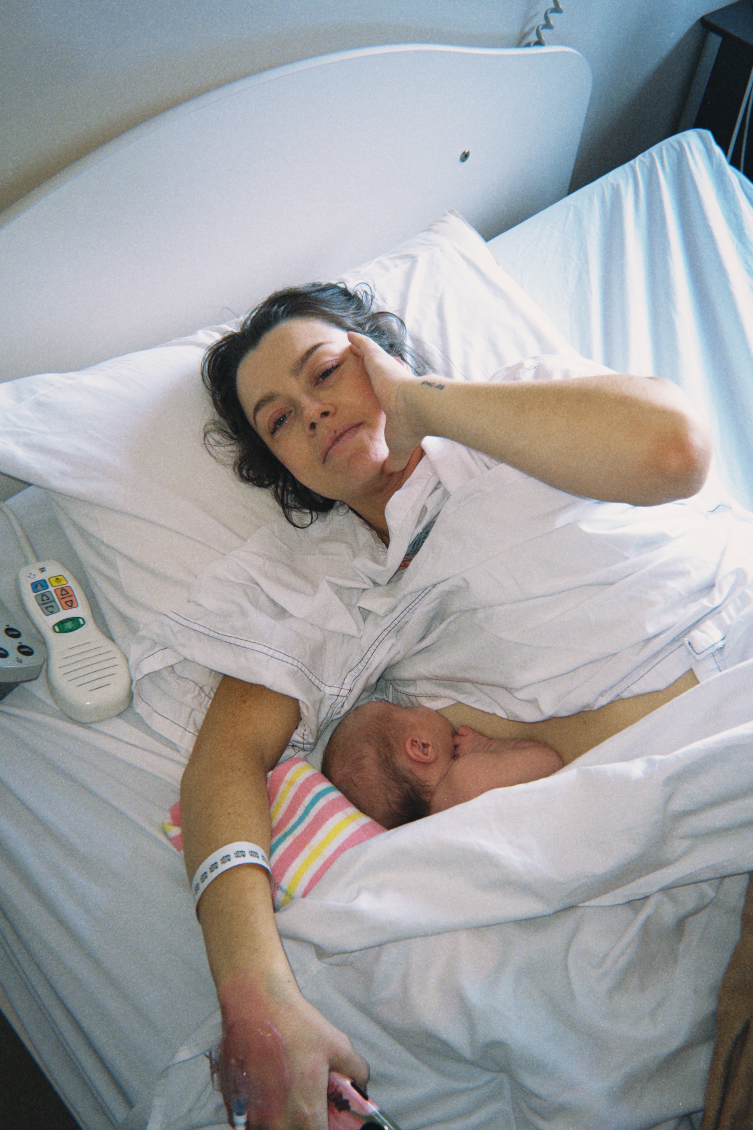 A tired-looking woman reclines on the white linen of a hospital bed while breastfeeding.