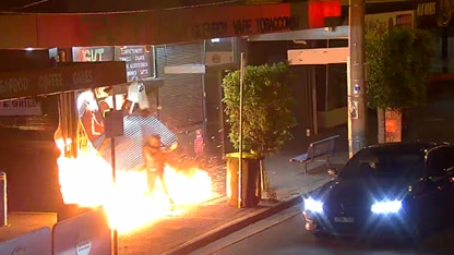 A man in the middle of flames engulfing the front of a shop, with a car parked on the road next to him, headlights on.